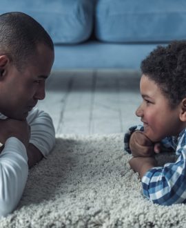 Happy Afro American father and son in casual clothes are looking at each other and smiling while lying on the rug at home